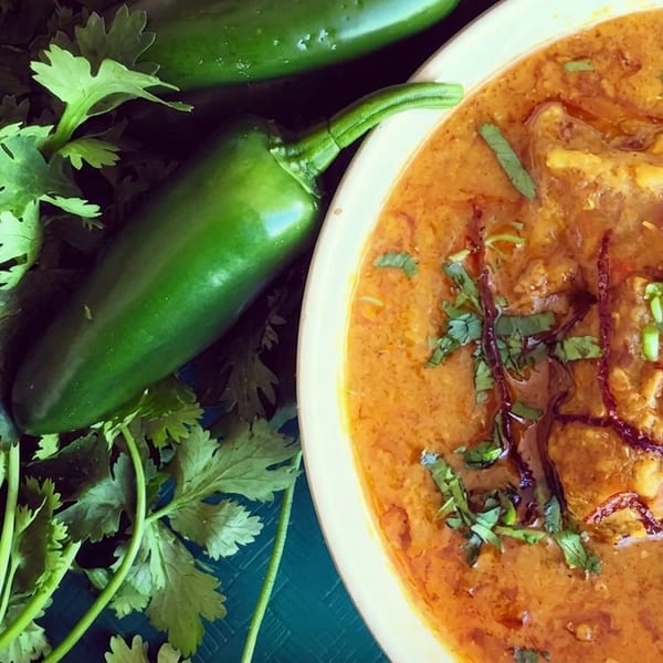 A bowl of haleem, garnished with fried onions and cilantro, next to fresh cilantro and jalapeños.