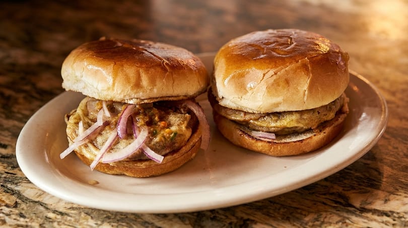 Two sliders with patties, sauce, and red onions on a white plate, on a marble surface.