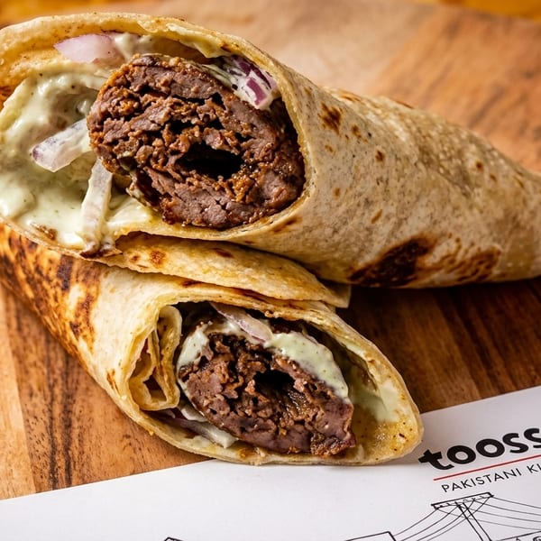 Two Toosso Pakistani Kitchen beef seekh kebab wraps with sauce and onions on a wooden board.