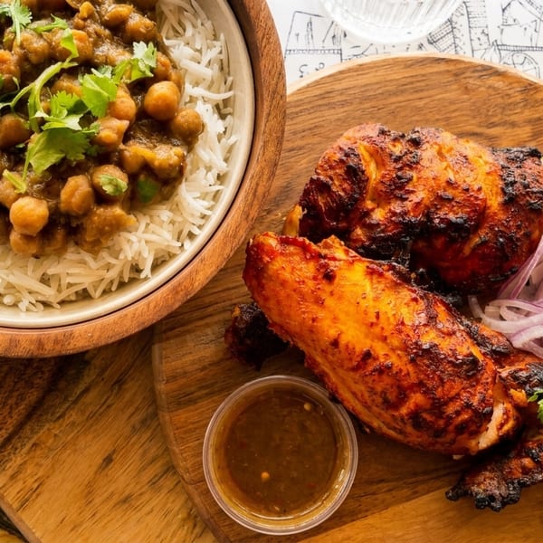 A bowl of chickpea curry with rice next to grilled chicken, red onions, and lemon wedges.