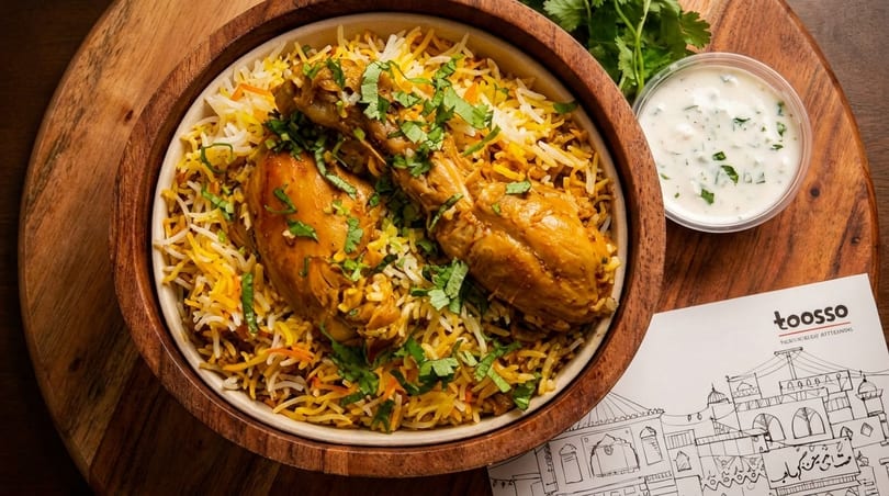 Chicken biryani with two chicken pieces, rice, cilantro, and a side of raita on a wooden board.